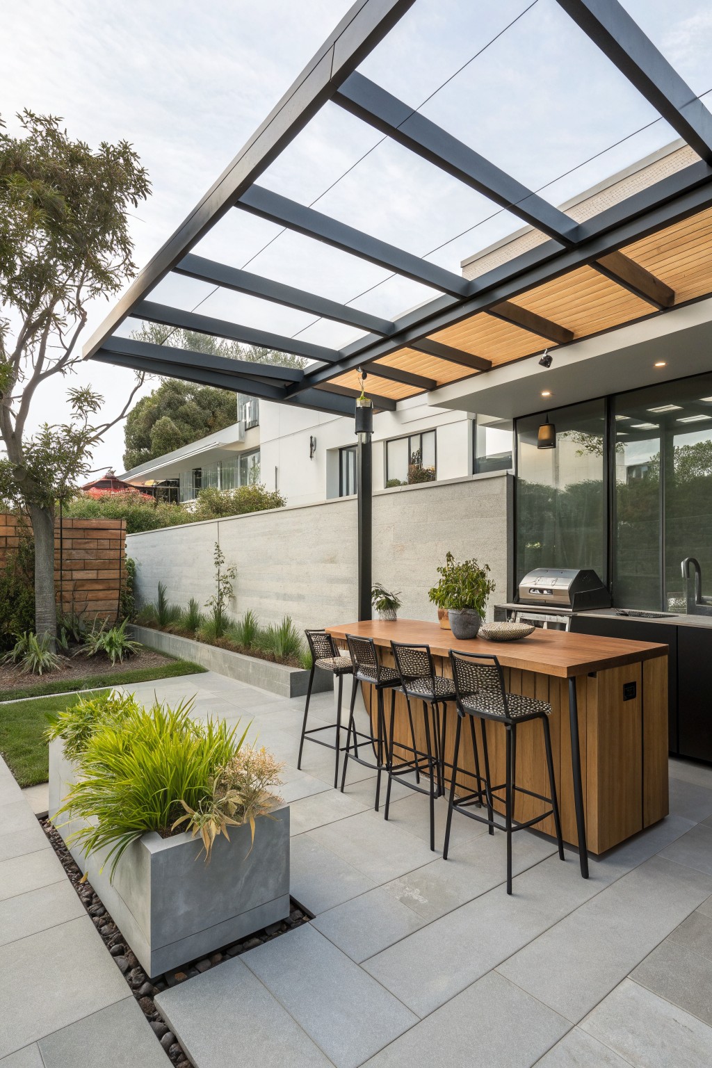Modern outdoor kitchen with wooden island, four bar stools, grill under glass roof.