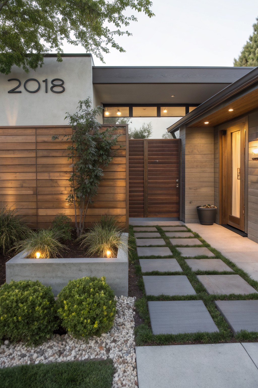 A modern house front with a tall wooden privacy fence of horizontal slats, a matching gate, a concrete planter with small trees, and a stone paver path leading to the entry door.