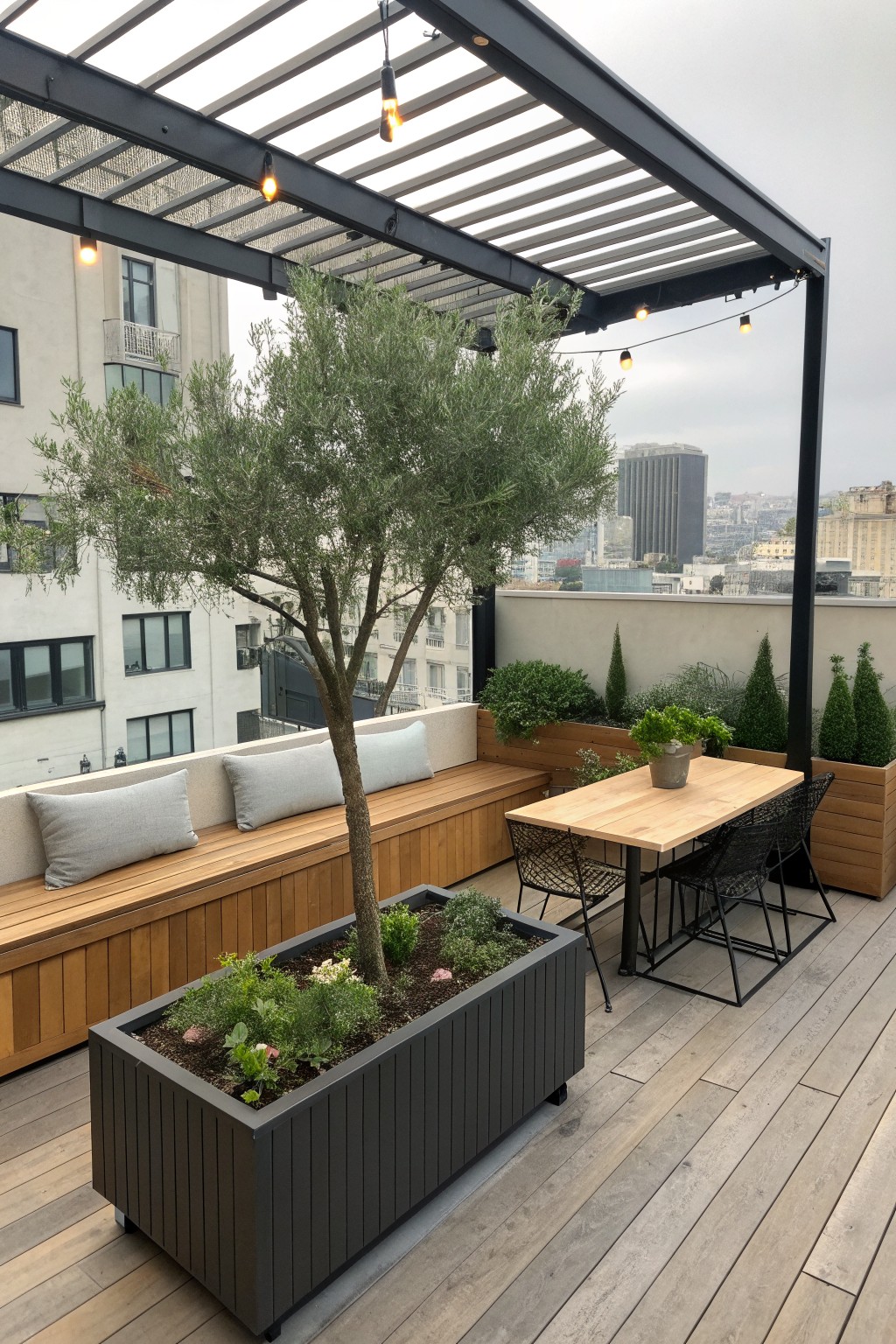 A rooftop terrace shows a built-in wooden bench with gray cushions beside a large gray planter holding a small tree, with a wooden dining table and chairs nearby under a black pergola.