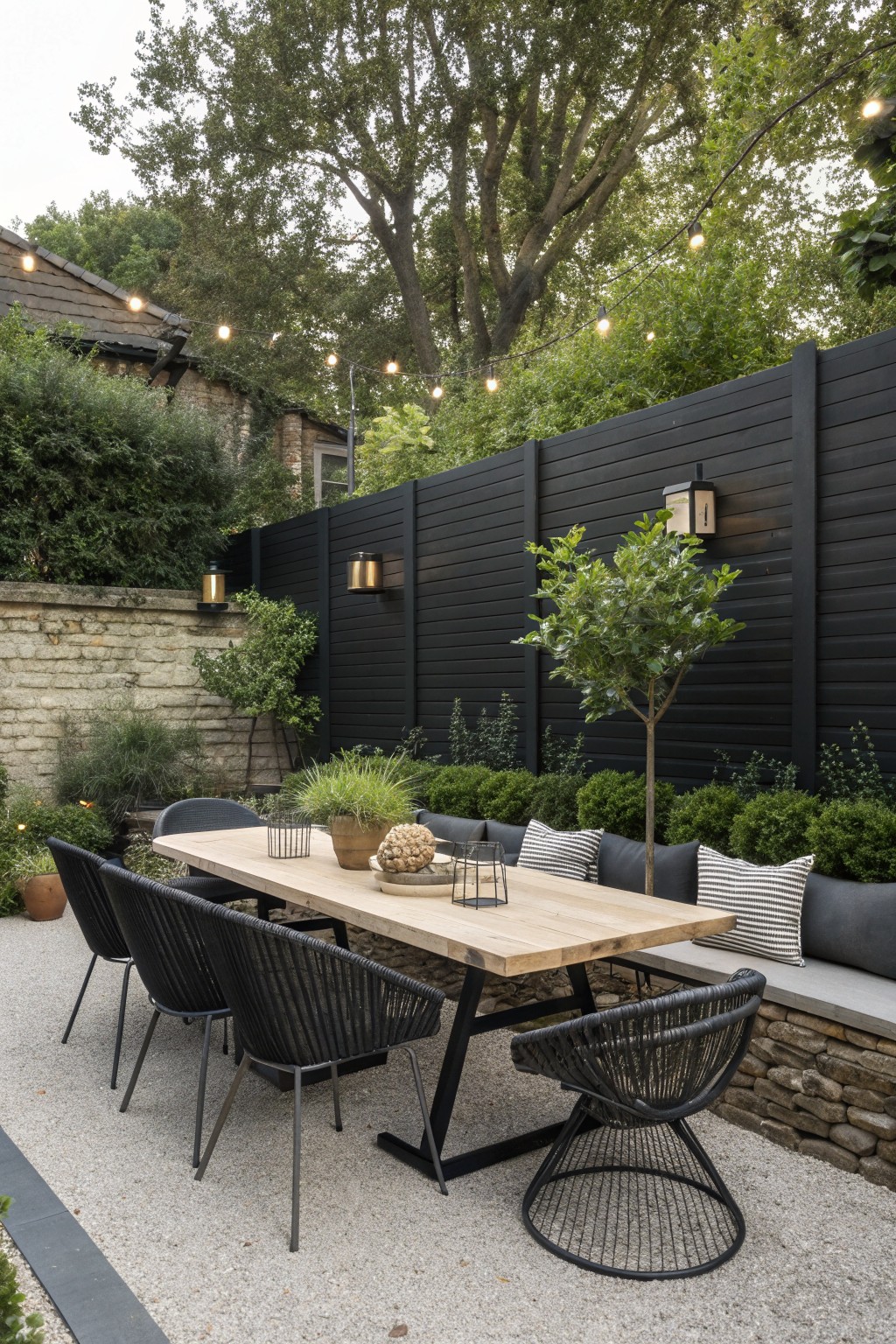 Outdoor dining setup with a light wood table, black chairs, and built-in bench seating in front of a tall black horizontal slatted fence.