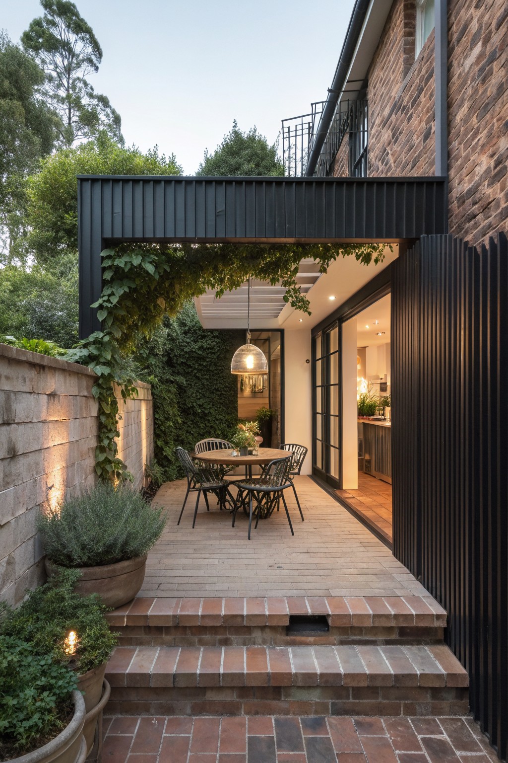 Outdoor patio with round table and chairs beneath a black slatted overhead structure, bordered by vertical black slats on the right and brick steps in the foreground.
