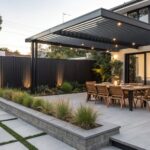 Modern outdoor patio with a long wooden dining table under a black metal slatted pergola, bordered by a tall black vertical slat fence and concrete planters with ornamental grasses.