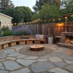 A curved stone wall topped with wooden benches in a backyard patio, surrounded by potted plants, a metal fire pit, string lights, and a stone path.