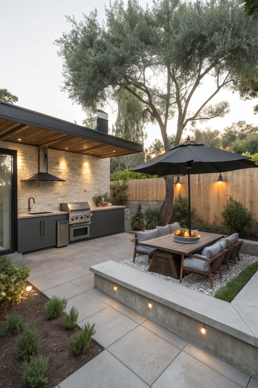 A modern outdoor patio with a built-in concrete seating ledge, wooden dining table under a black umbrella, and an outdoor kitchen area against a stone wall.
