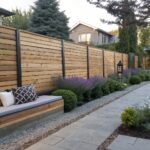 A long horizontal cedar fence runs beside a gravel path with stone pavers, a wooden bench with cushions, and a border of lavender and green shrubs.