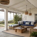 A covered patio shows a long wooden sofa with blue striped cushions and pillows, a wooden coffee table on a rug, and woven pendant lights hanging from exposed beams.