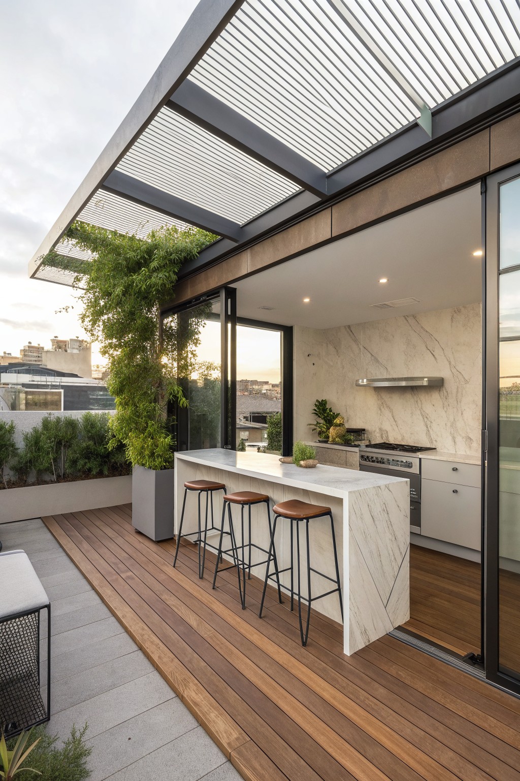 Rooftop outdoor kitchen with a white marble bar counter, three bar stools, and a dark slatted pergola overhead on a wooden deck.