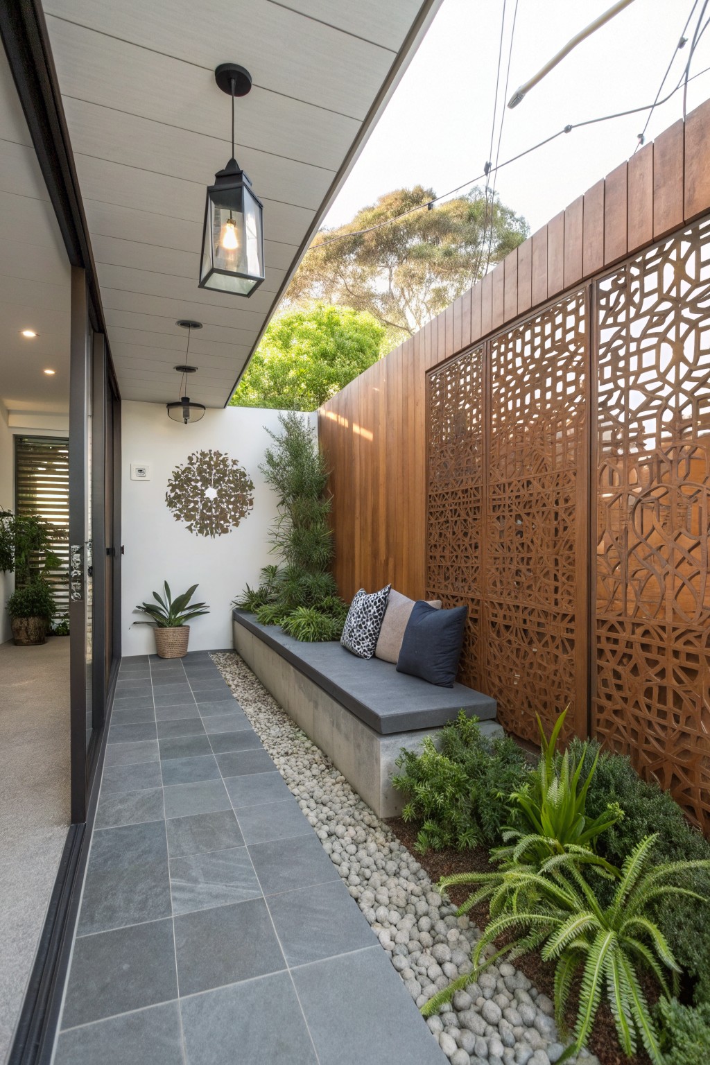 A narrow outdoor patio with gray tiles, a built-in bench with cushions, plants along the edge, and a wooden fence with decorative cutout panels.