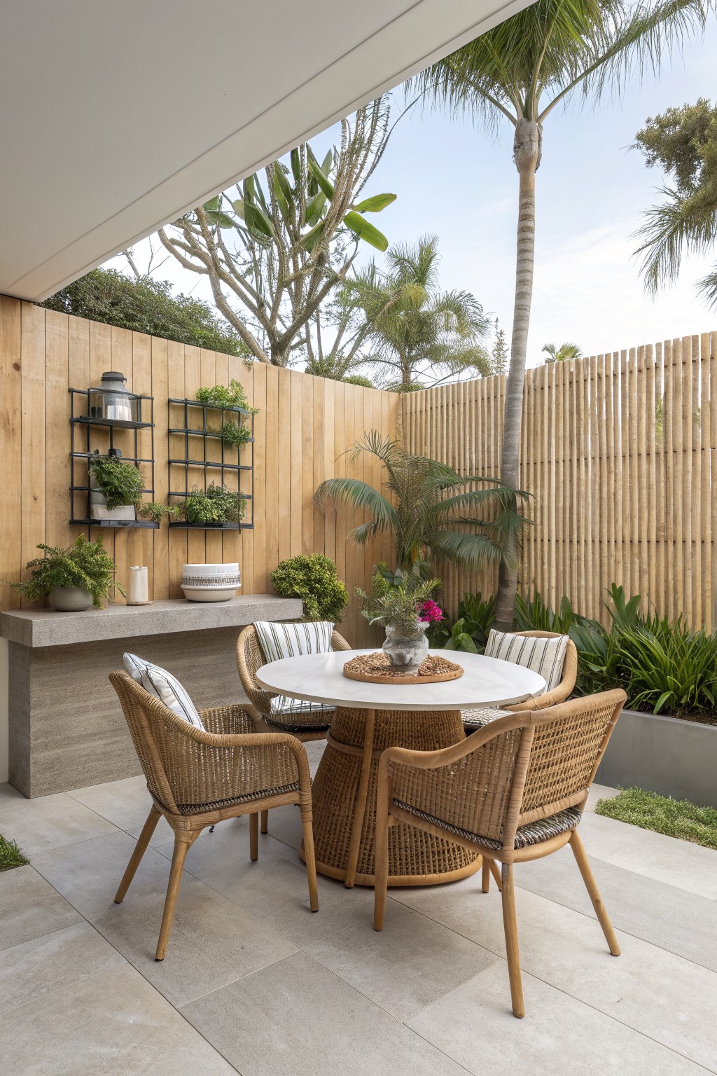 An outdoor patio with a round table and wicker chairs sits in front of a tall vertical bamboo privacy fence surrounded by tropical plants.