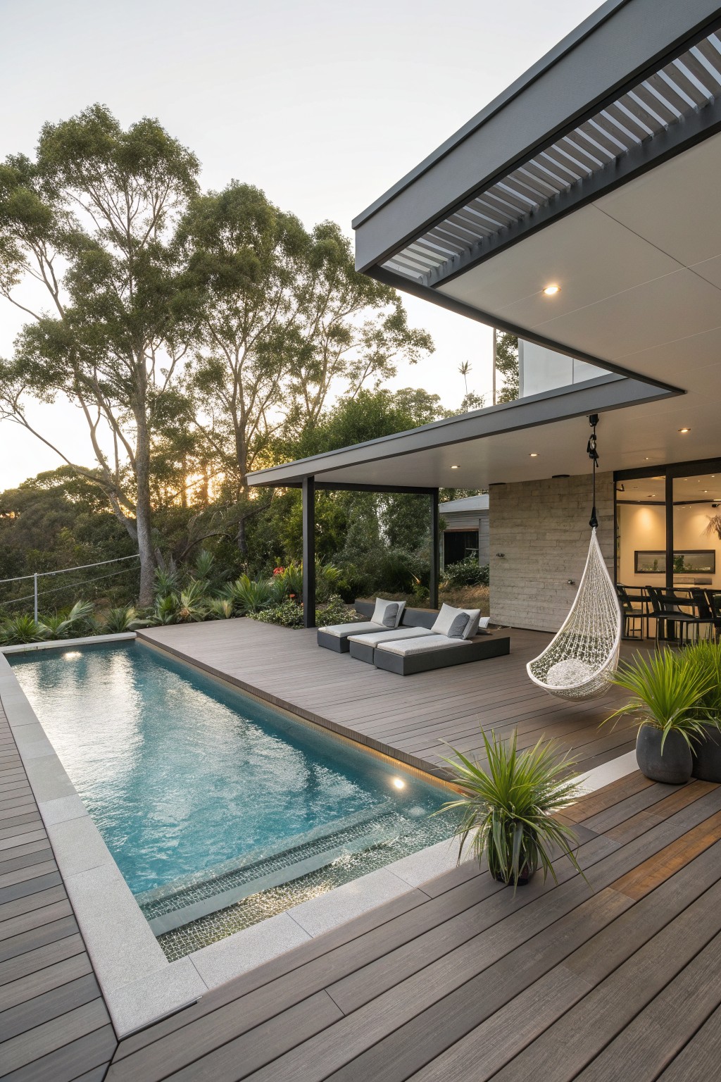 Modern poolside deck with lounge sofas, hanging chair, potted plants, and sunset view.