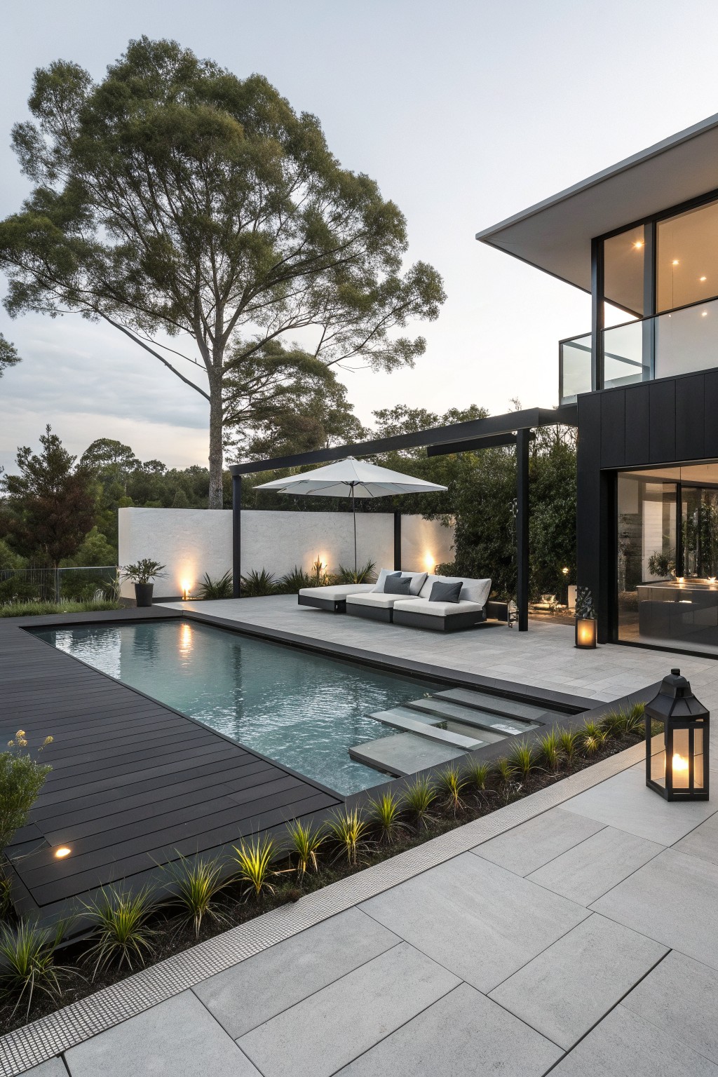 A modern outdoor pool area with a rectangular swimming pool, dark decking, a light gray sectional sofa under a black pergola with a white umbrella, and a contemporary house with large glass doors on the right.
