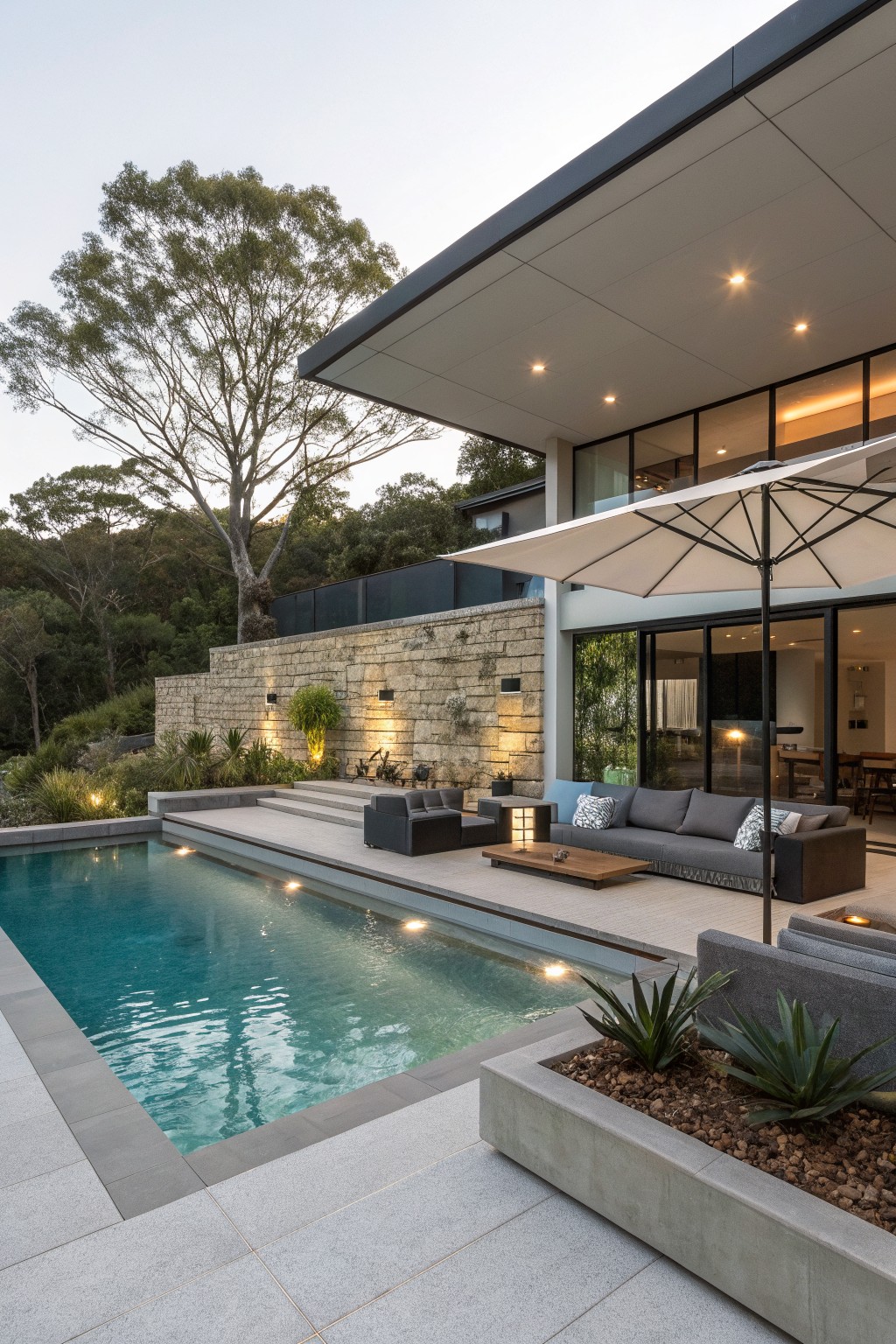 A modern outdoor pool area with a long rectangular pool and gray sectional seating arranged on a paved patio under a large white umbrella.