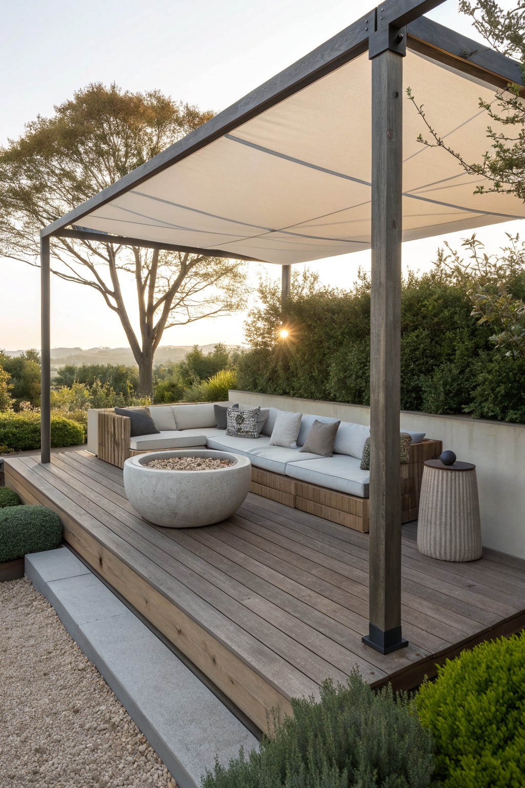 Wooden deck with built-in low-profile sofas arranged around a round concrete fire pit beneath a fabric shade pergola, bordered by plants and trees.