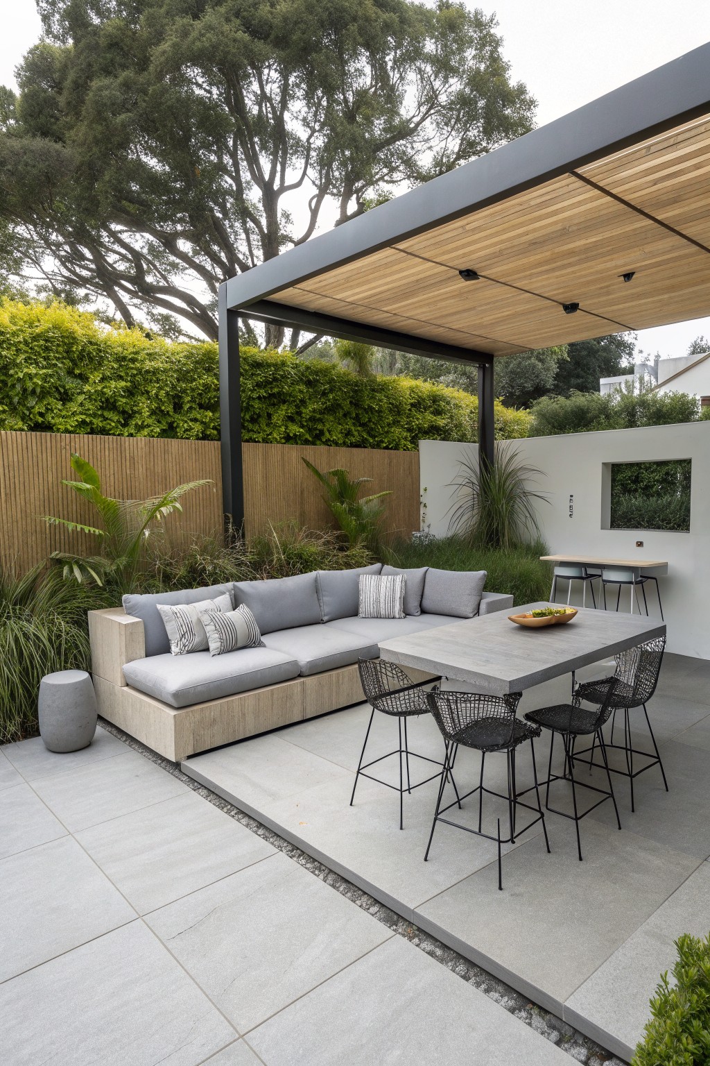 A covered outdoor patio features a low-profile gray sectional sofa with striped pillows beneath a dark metal pergola with wood slats, next to a concrete dining table and black wire bar stools.